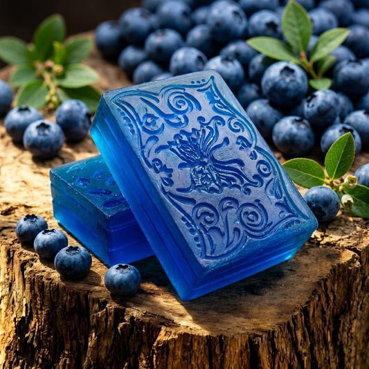 Two translucent blue rectangular bars of soap with embossed floral and scroll patterns, stacked on a rustic wood slice and surrounded by fresh blueberries and green leaves. Made in Canada with glycerin soap base and paraben and phthalates free oils. $6.00 CAD