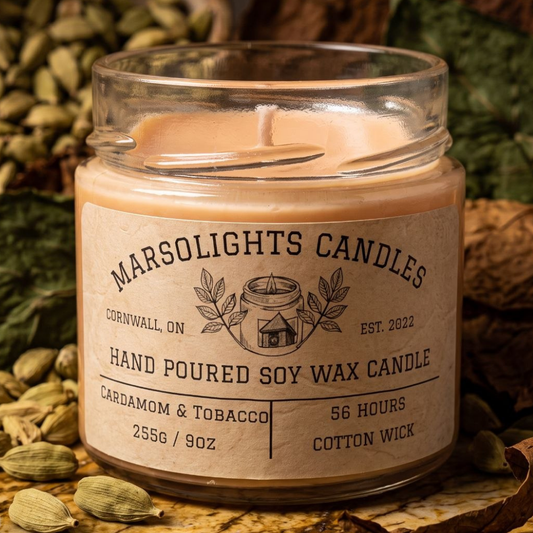Cardamom & Tobacco Soy Candle on a table with warm, sophisticated décor. Our candle features notes of cardamom, tobacco leaf, jasmine, cashmere musk, vanilla, cedarwood, cinnamon, clove, and tonka bean. 9 ounce or 255 grams, 56-hour burn time, cotton wick, freedom soy wax, made in Canada, paraben free.