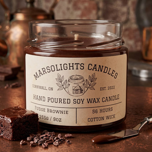 Fudge Brownie Soy Candle displayed with chocolate brownie-inspired décor. The candle features rich chocolate fudge, smooth milk chocolate and toasted walnut notes. 9 ounce or 255 grams, 56-hour burn time, cotton wick, soy candle, made in Canada, paraben free.